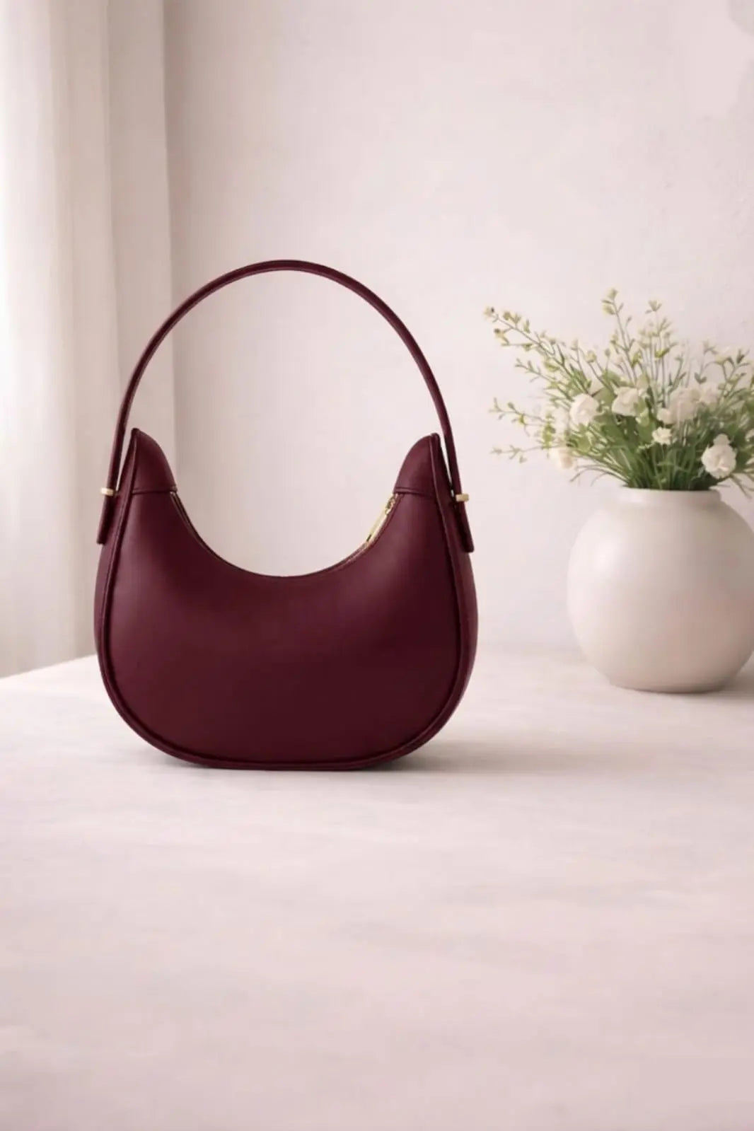 A crescent-shaped leather shoulder bag in deep burgundy with gold-tone hardware, displayed on a light beige surface next to a white ceramic vase with small white flowers.
